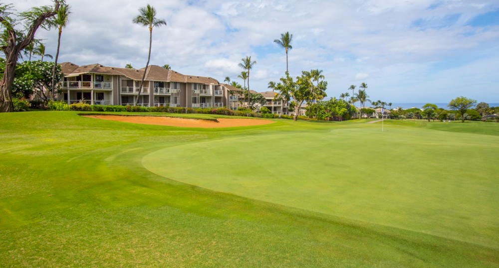 A lush golf course with neatly trimmed greens and a sand bunker, adjacent to modern, tropical-style buildings with palm trees on a bright day.