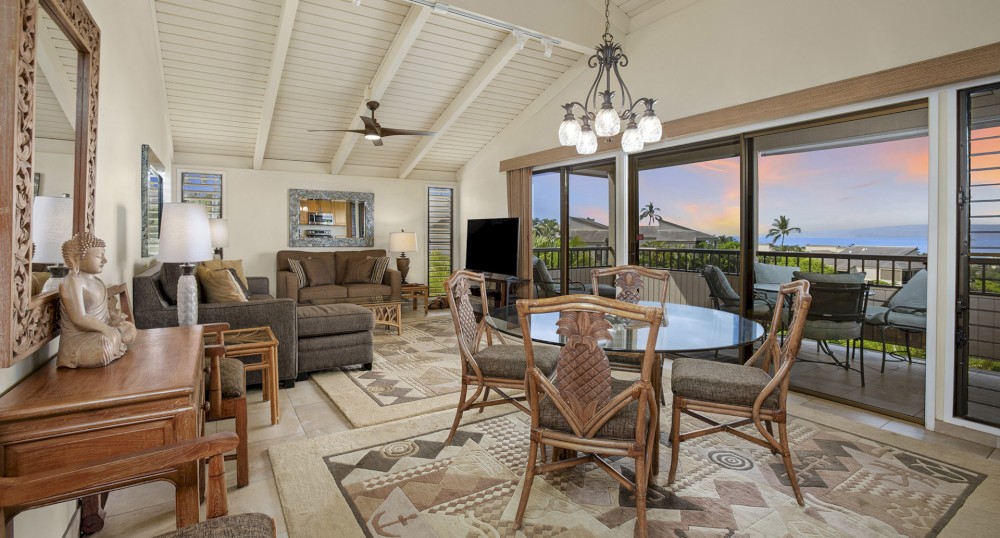 A cozy living space featuring a sectional sofa, a wooden table with chairs, art pieces, and a view of the ocean visible through large windows.