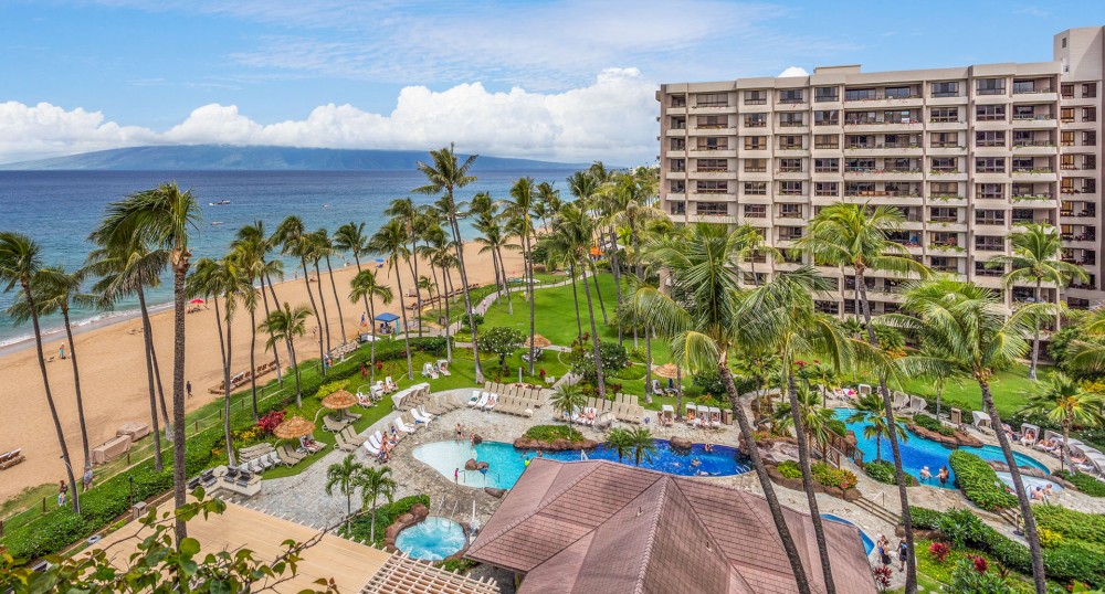 A beachfront resort with palm trees, swimming pools, lounge chairs, and a multi-story building overlooking the ocean on a sunny day.