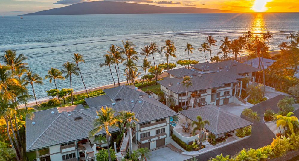An oceanfront property with palm trees, houses, and a beautiful sunset over the sea. The landscape features a peaceful and picturesque setting.