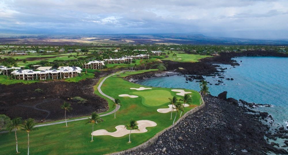 A scenic coastal golf course with green fairways, sand traps, palm trees, and nearby buildings overlooks a rocky shoreline under a cloudy sky.