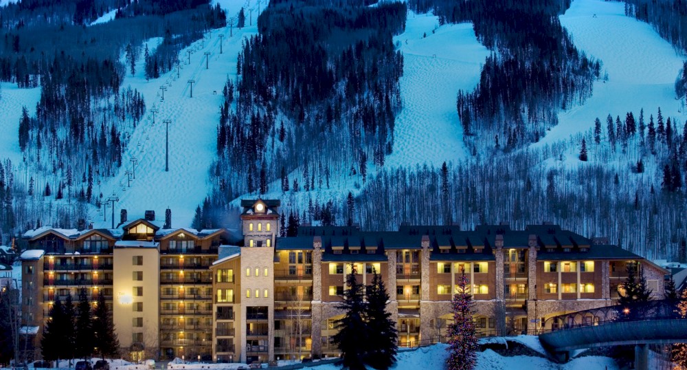 A well-lit resort nestled at the base of a snowy mountain with ski slopes and a dense forest of pine trees in the background, captured at dusk.