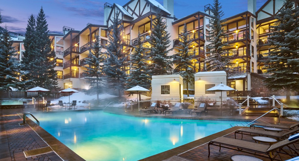 The image shows a luxurious resort with an outdoor heated pool surrounded by lounge chairs and umbrellas. Snow covers the nearby trees and buildings.