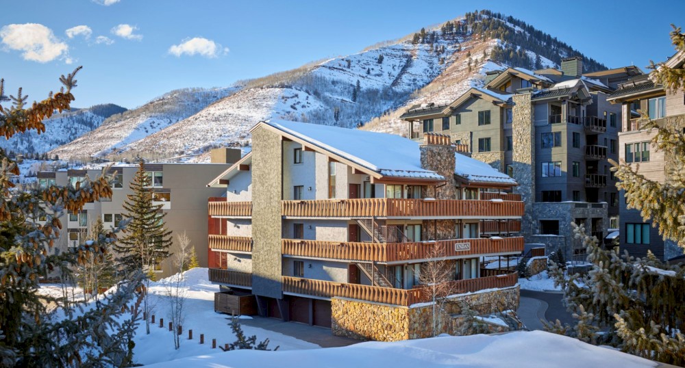 The image shows a snow-covered mountainous area with multiple buildings, including a wooden lodge, surrounded by pine trees and a clear blue sky.