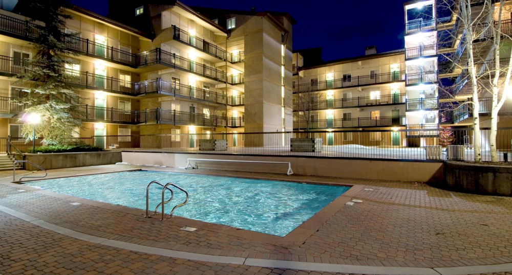 An illuminated apartment complex at night with an empty outdoor swimming pool, surrounded by a paved patio area and railings.