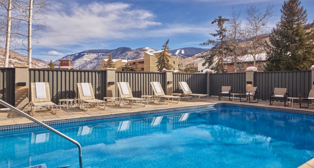 An outdoor pool with lounge chairs and a mountainous backdrop framed by trees and a clear sky in the background.