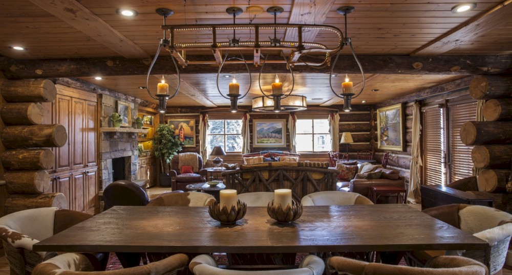 A cozy, rustic living room with a wooden dining table, log walls, a chandelier with candles, cushioned chairs, and large windows.
