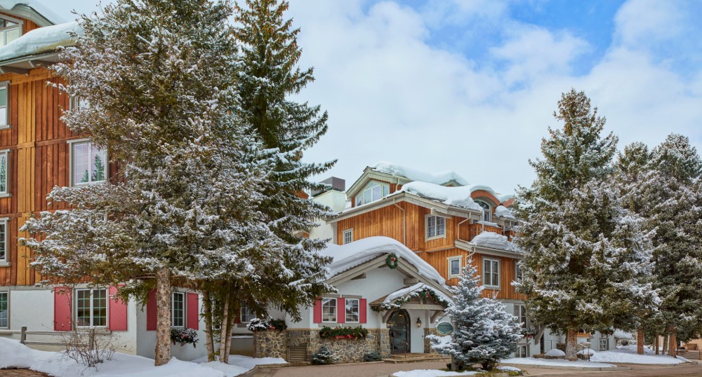 A cozy wooden building surrounded by snow-covered pine trees under a partly cloudy sky. The scene has a winter wonderland feel to it.