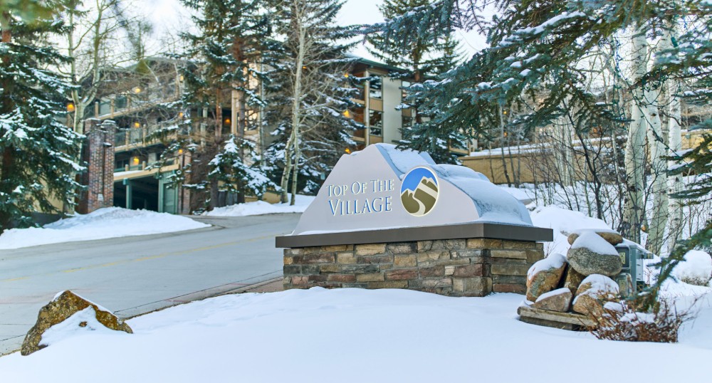 A snow-covered entrance sign reads "Top of the Village," with buildings and trees in the background.
