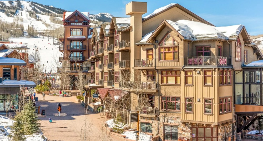 A picturesque ski resort with snow-covered buildings and mountains in the background. People walk along the paths, enjoying the sunny day.
