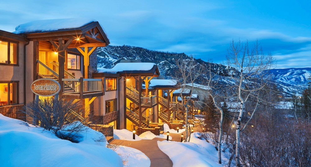 A snow-covered lodge with warmly lit buildings and a scenic mountain backdrop during twilight, creating a cozy and inviting atmosphere.