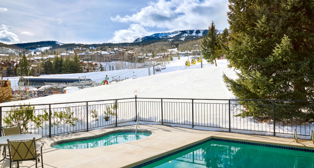 The image shows a pool and hot tub with a snowy mountain backdrop, featuring chairs and a fence, under a partly cloudy sky.