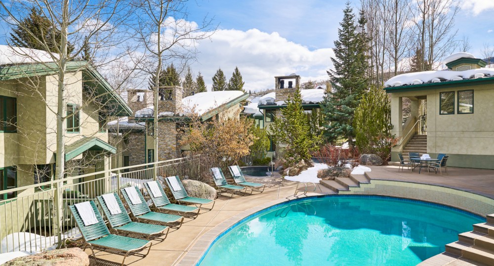 This image features a serene outdoor pool area surrounded by lounge chairs and apartment buildings set against a snowy, wooded backdrop.