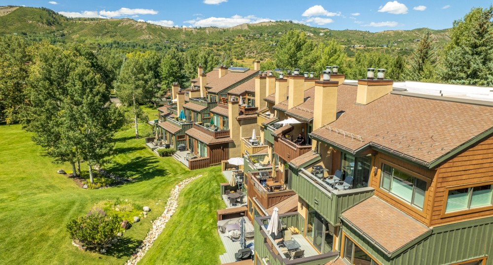 A row of modern, multi-story townhouses situated in a lush, green environment with ample trees and a scenic mountain backdrop under a bright blue sky.