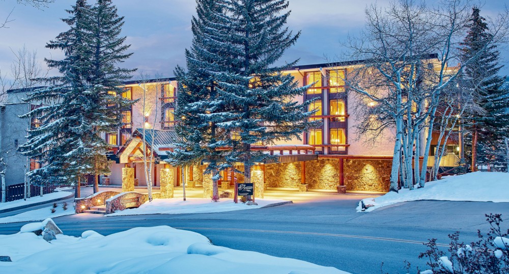 The image shows a well-lit, cozy building surrounded by snow, trees, and a neatly plowed road, possibly a hotel or lodge during winter.