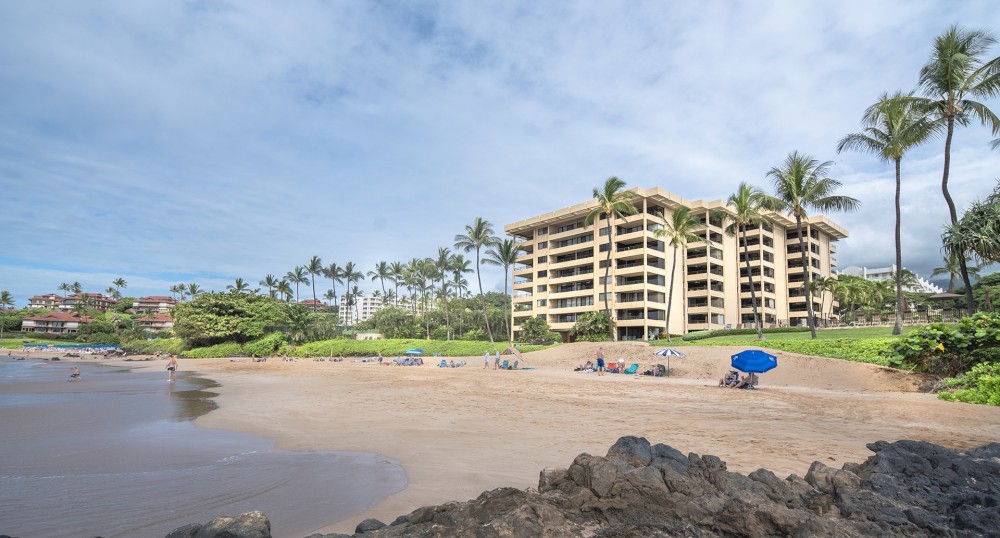 A beach scene with people relaxing on the sand, umbrellas, a sandy shore, palm trees, and a multi-story building in the background.