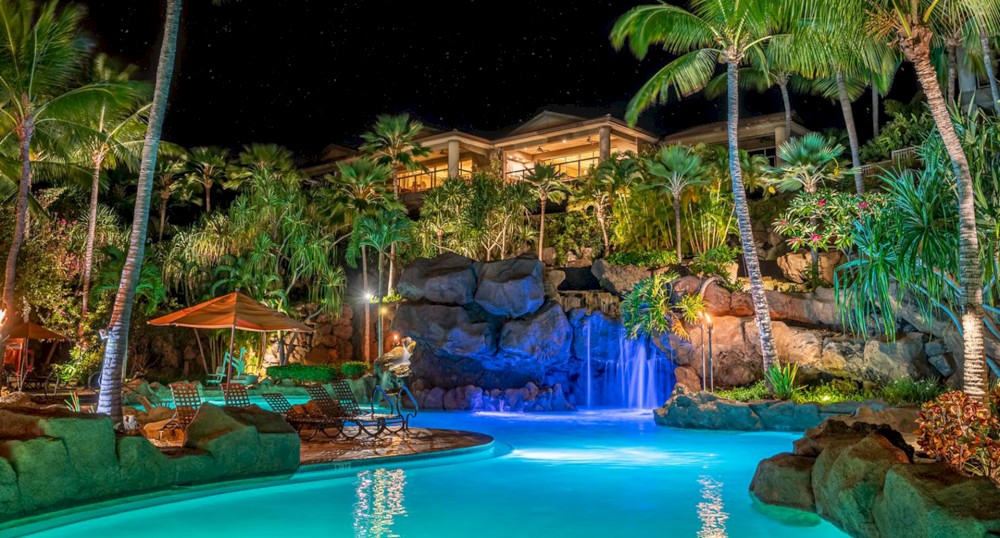 A vibrant nighttime view of a tropical resort, featuring a glowing pool, a waterfall, lush palm trees, and lounge chairs under an umbrella.