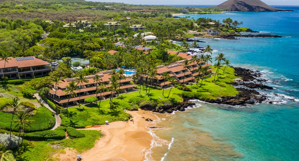 Aerial view of a resort along a shoreline with clear blue waters, sandy beach, and lush green surroundings on a tropical island.