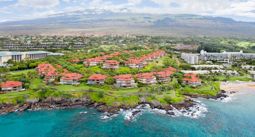 An aerial view shows red-roofed buildings along a scenic coastline with blue waters, a grassy landscape, and mountains in the background.