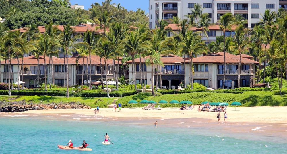 The image shows a tropical beach scene with people kayaking and lounging under umbrellas, flanked by lush palm trees and luxury buildings in the background.
