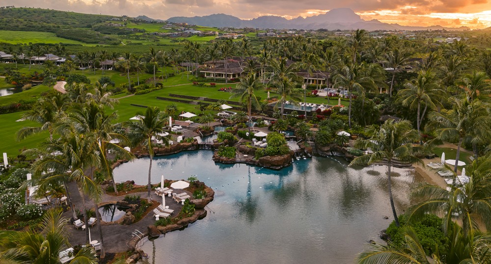 The image depicts a serene resort with a large pool surrounded by lush greenery, palm trees, and various small buildings, set against a mountainous backdrop.