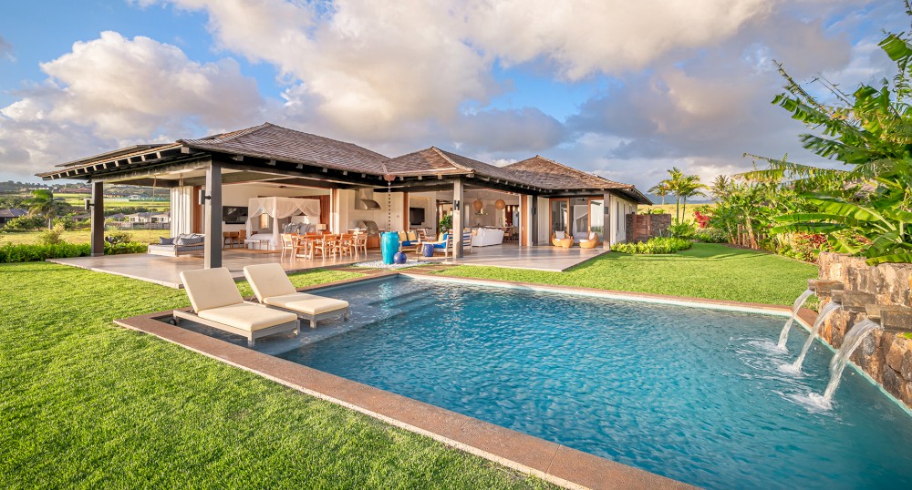 A luxurious villa with open living spaces, outdoor seating, lush lawn, and a swimming pool with lounge chairs in a tropical setting under a sunny sky.