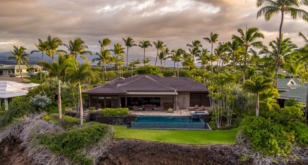 A beautiful tropical house with a pool is surrounded by palm trees, greenery, and a rocky terrain under an overcast sky.