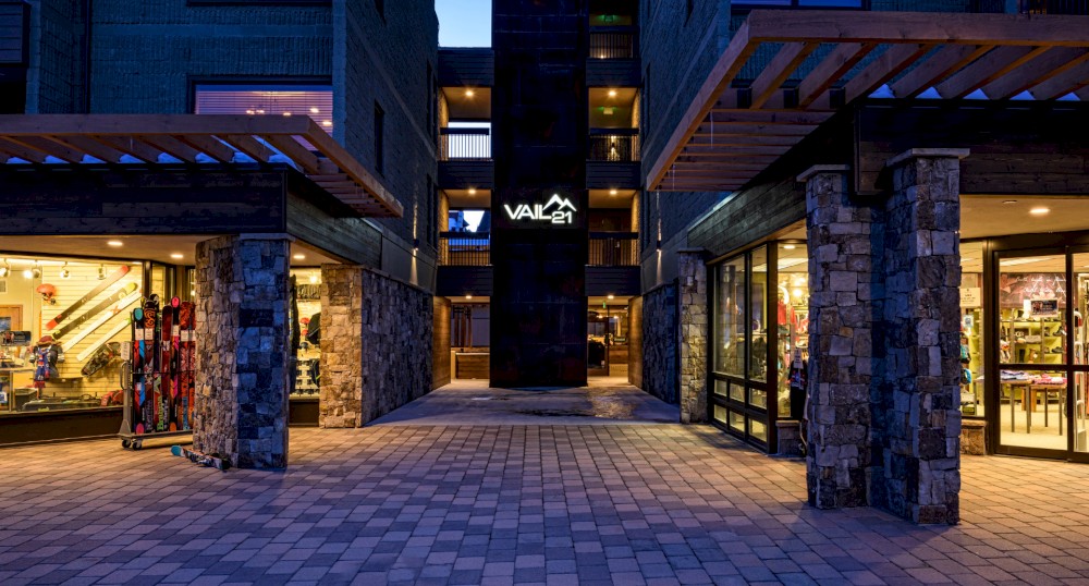 The image shows a well-lit commercial area at dusk with a sign that reads "VAIL." Stores with large glass windows display various items.