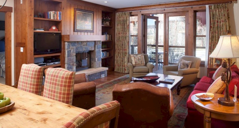 A cozy living room with wooden furniture, a stone fireplace, plaid chairs, a red sofa, and large windows with curtains leading to a balcony.