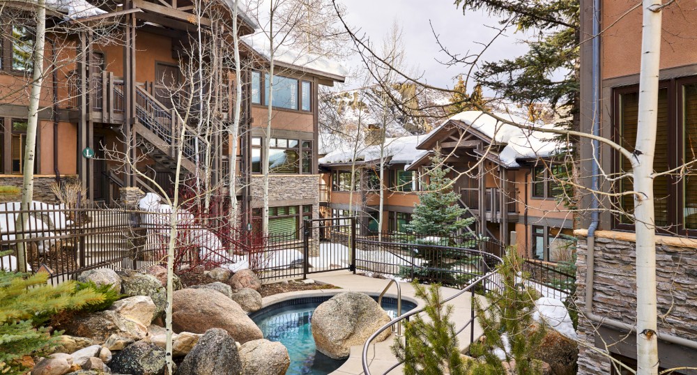 The image shows a cozy courtyard with rustic buildings, snow-covered rooftops, evergreen trees, and a small outdoor hot tub surrounded by rocks and a fence.