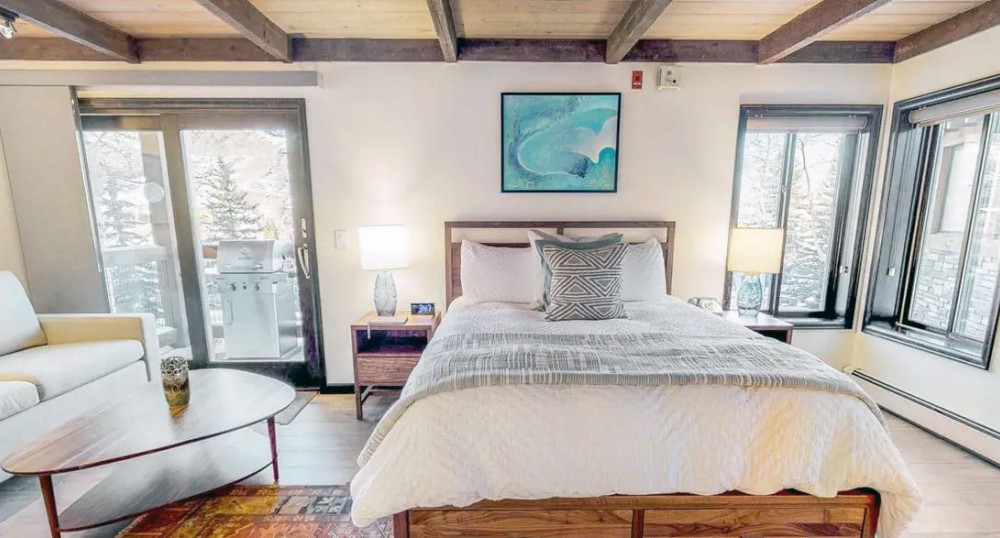 A cozy bedroom with a wooden beam ceiling, featuring a bed, sofa, coffee table, and art. Bright and inviting with large windows and a sliding door.