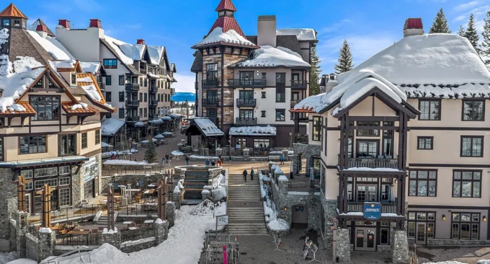 A snowy village with Alpine-style buildings, snow-covered roofs, and people walking in the central area on a clear, sunny day.