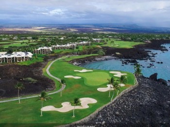 The image shows a golf course near the ocean, with green fairways, sand bunkers, and surrounding residential buildings amidst a scenic landscape.