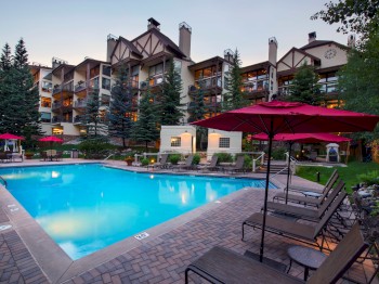 An outdoor pool with lounge chairs and red umbrellas at a resort or hotel, surrounded by multi-story buildings and tall pine trees.