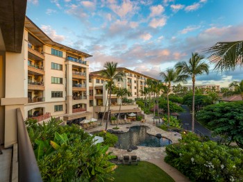 A tropical resort with multi-story buildings, lush greenery, and a pool area surrounded by palm trees under a partly cloudy sky.